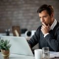 Smiling businessman using laptop and contemplating while working at his desk in the office.